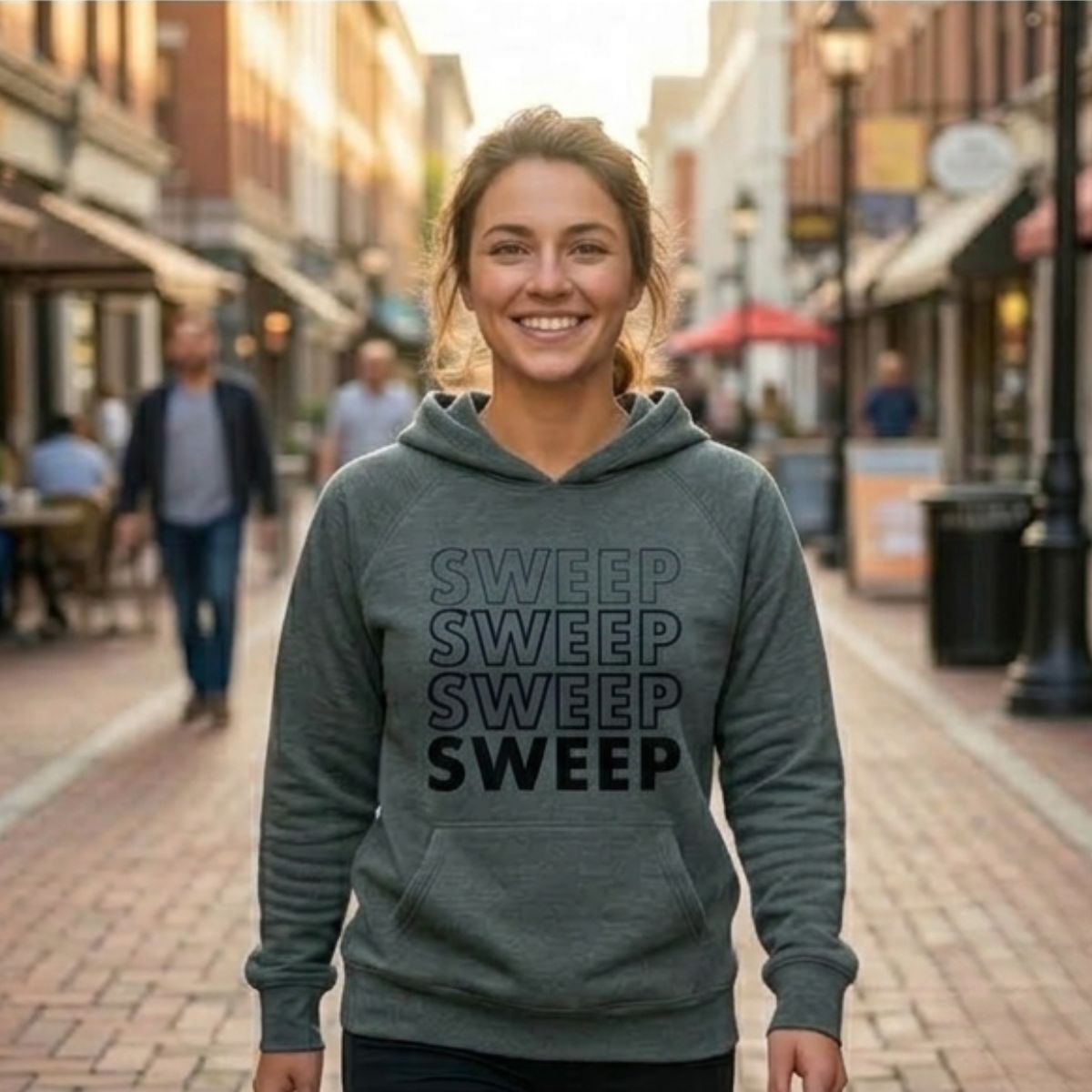 A side-by-side comparison of a woman walking down a charming, sunlit brick street. On the left, she is wearing the "SWEEP" hoodie in Charcoal Grey; on the right, she wears the same design in Indigo Blue. Both hoodies feature a bold, vertical stack of the word "SWEEP" in a gradient-style print.