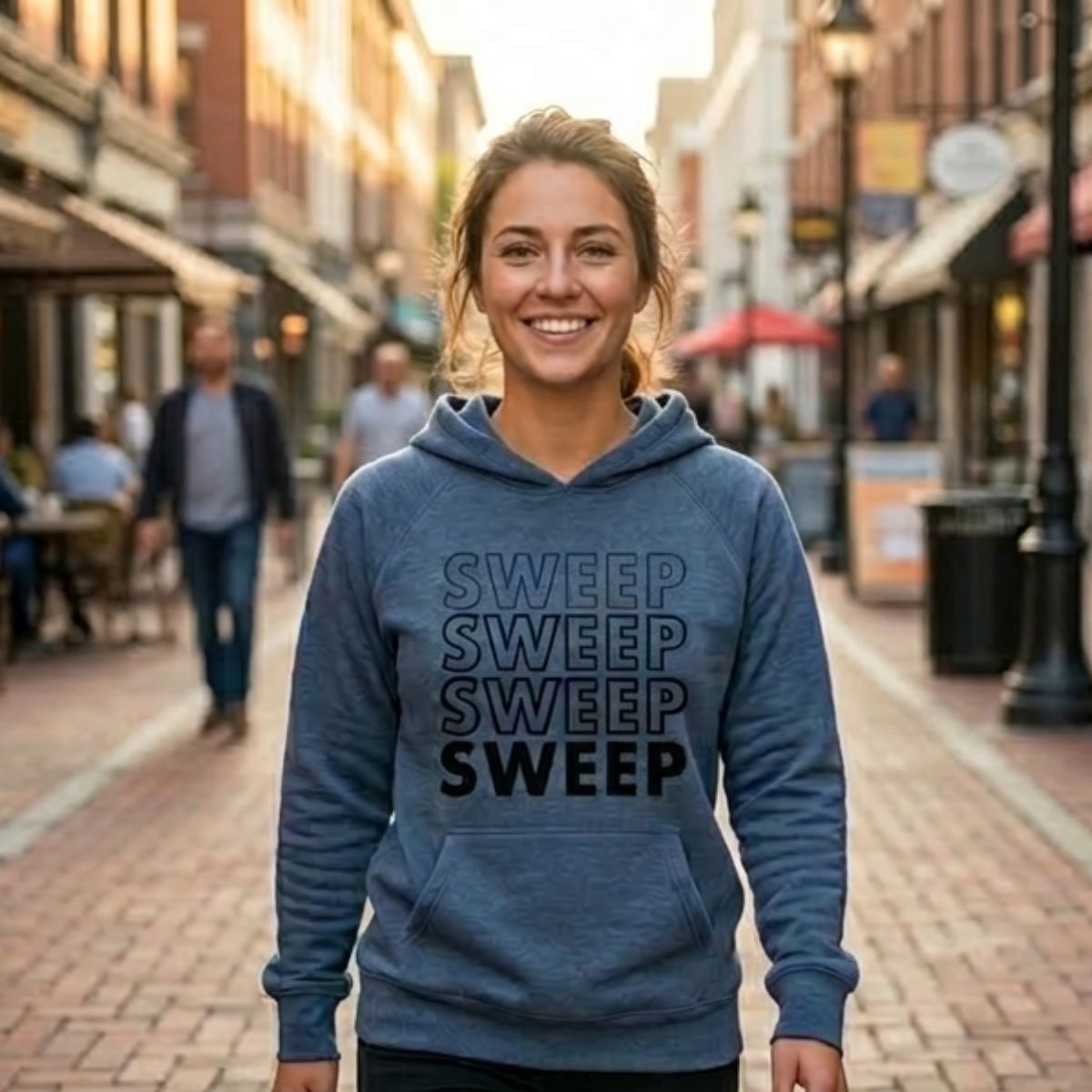 A side-by-side comparison of a woman walking down a charming, sunlit brick street. On the left, she is wearing the "SWEEP" hoodie in Charcoal Grey; on the right, she wears the same design in Indigo Blue. Both hoodies feature a bold, vertical stack of the word "SWEEP" in a gradient-style print.