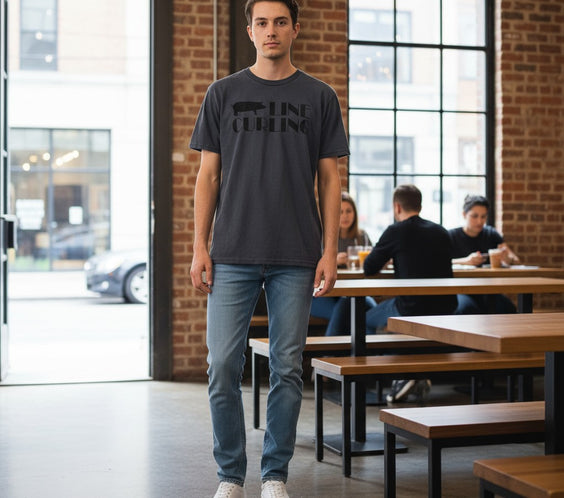 A man standing in a modern cafe wearing a dark gray Hog Line Curling T-shirt with a hog silhouette and bold text across the chest. He is dressed casually in jeans and white sneakers, with brick walls and large windows in the background.