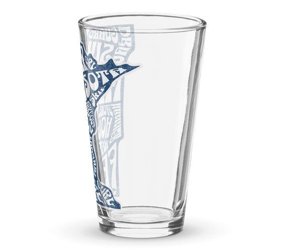 Angled view of a clear shaker pint glass featuring a blue Minnesota state outline with curling terms such as sweep, guard, button, freeze, and hog line, partially visible along the side of the glass. Curling-themed drinkware by CRL.