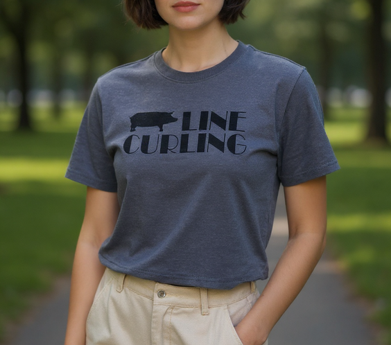 A woman standing on a park path wearing a dark gray Hog Line Curling T-shirt featuring a hog silhouette and bold lettering. She is dressed casually in light-colored pants, with trees and greenery blurred in the background.