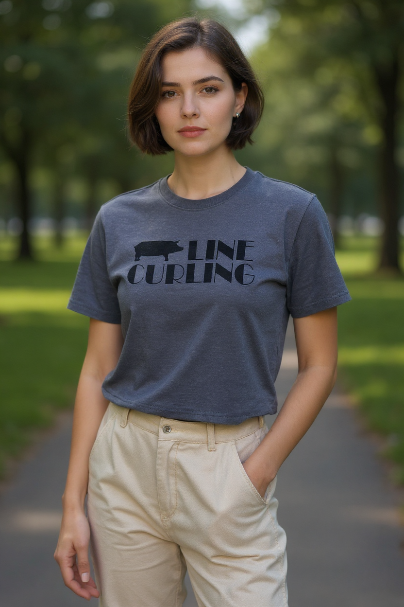 A woman standing on a park path wearing a dark gray Hog Line Curling T-shirt featuring a hog silhouette and bold lettering. She is dressed casually in light-colored pants, with trees and greenery blurred in the background.